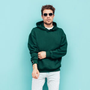 handsome confident hipster model.sexy unshaven man dressed in summer stylish green hoodie and jeans clothes. fashion male with curly hairstyle posing in studio. isolated on blue