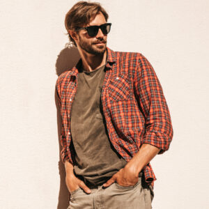 portrait of handsome stylish hipster lambersexual model.man dressed in checkered shirt. fashion male posing in the street near white wall outdoors in sunglasses